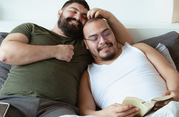 Gay plus sized male couple smiling while reading in bed