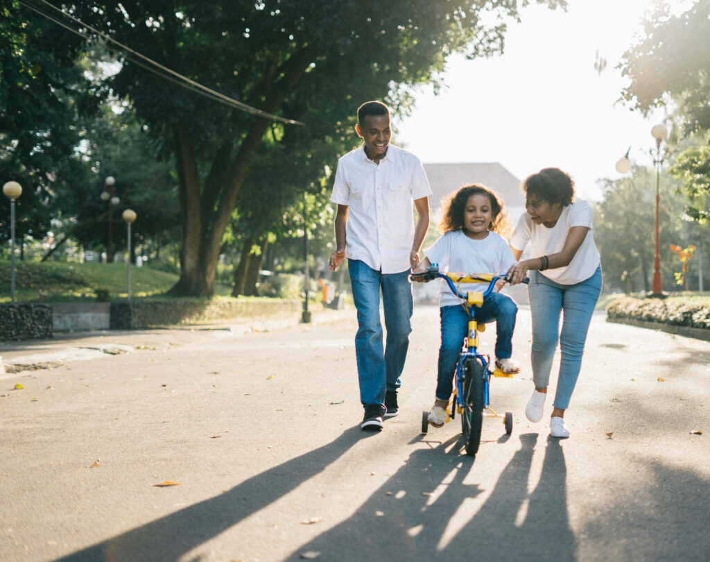 Black Gen Z couple teaching their young daughter to ride her bike on a street at sunset